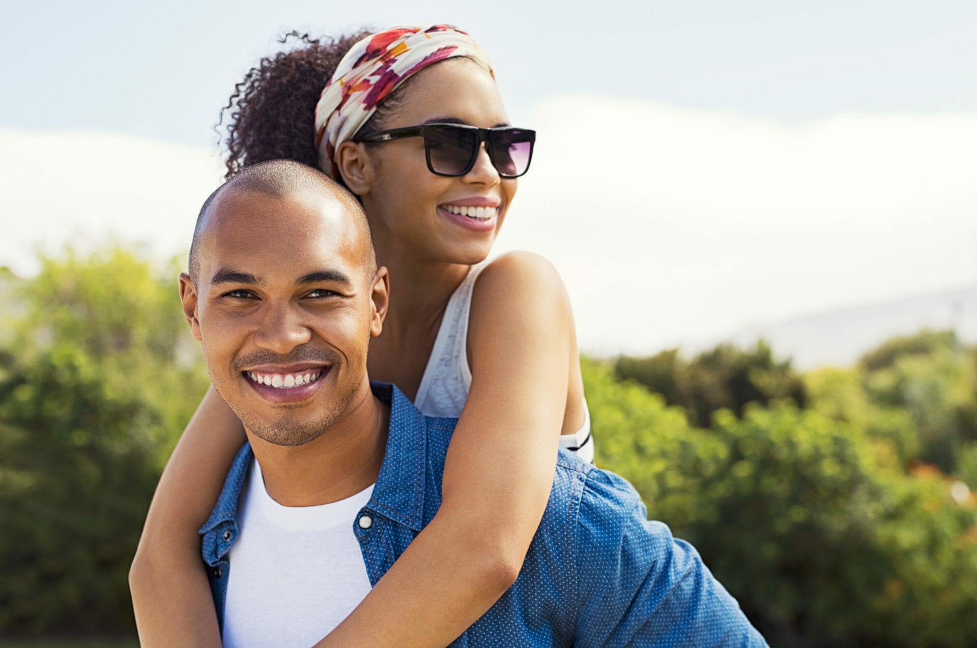Portrait of young black man holding beautiful woman on back and looking at camera. African american woman with sunglasses enjoying piggyback on boyfriend at park while looking away. Smiling couple enjoying the summer.